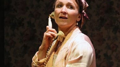 A woman in a pink dressing gown and headscarf stands indoors, holding a vintage cream-coloured telephone to her ear and looking upward with a thoughtful expression. A floral wall and a handbag are visible in the background.
