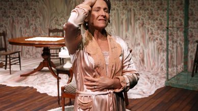 A woman in a satin dressing gown stands with one hand on her head and the other on her chest, eyes closed, in a vintage-style room with wooden floors, floral wallpaper, and antique furniture.