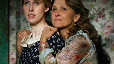 A woman in a floral blouse embraces a younger woman in a dark dress with a white collar and flower print, standing in front of a wall with floral wallpaper, both looking intently ahead.
