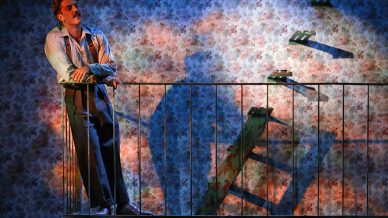 A man stands on an outdoor metal staircase, looking thoughtful, whilst a woman sits below him on a lower level. Dramatic lighting creates shadows on the floral wallpapered wall behind them.