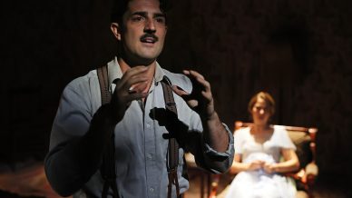 A man with a moustache, wearing braces and a button-up shirt, gestures expressively in the foreground. A woman in a light dress sits in a chair in the dimly lit background.