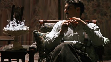 A man sitting in a dimly lit room on a cushioned chair looks to his right. A glass animal figurine centrepiece is on a cake stand on a nearby table. The atmosphere is moody and contemplative.