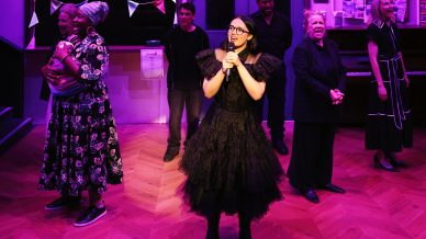A woman in a black dress sings into a microphone on stage, surrounded by five people in various outfits. The background includes bunting, posters, and wood-patterned flooring under purple stage lighting.