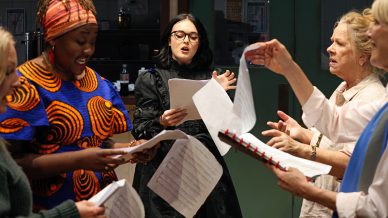 Five women stand in a circle holding sheet music, singing passionately together. One wears a vibrant, patterned dress and headwrap, another wears black glasses and dark clothing, while others are dressed in light-coloured, casual outfits.