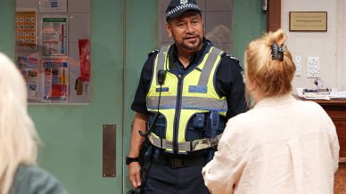 A police officer in uniform and high-visibility vest stands indoors, facing a woman with blonde hair who has her back to the camera. The setting appears to be an office or public building.