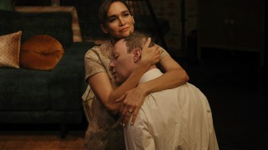 A woman sits on a sofa, gently embracing a man who kneels and rests his head on her chest. Her expression is thoughtful, while the man appears vulnerable. The room is dimly lit, with a green settee in the background.