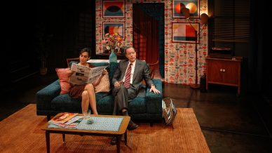 A woman reading a newspaper sits on a teal sofa next to a man in a suit and tie in a retro-style living room, with patterned wallpaper, colourful art, a coffee table, and a vintage record player in the background.