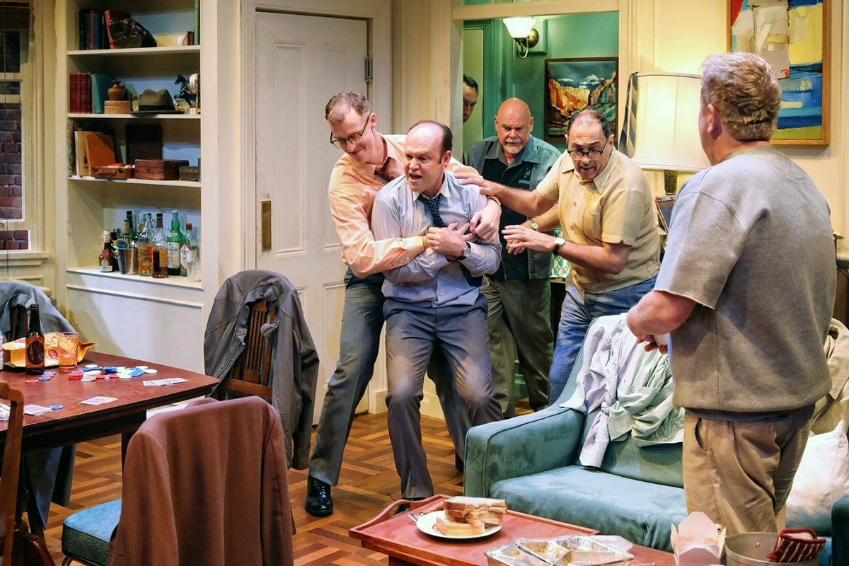 Five men are in a cluttered living room; three are restraining or supporting a distressed man in a suit, while another man stands nearby. The scene looks tense, with food and cards scattered on the table.