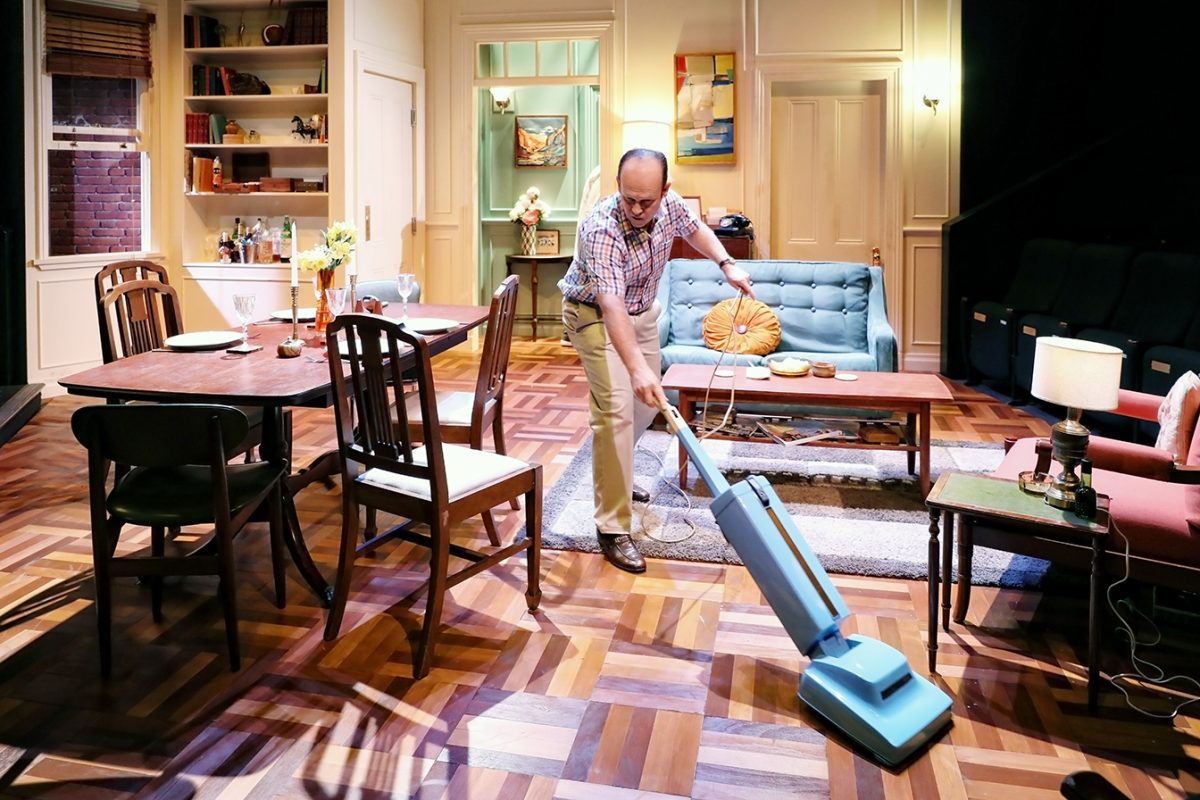 A man hoovers the floor in a cosy, well-lit living room with wooden furniture, a blue sofa, and various decorations, giving the scene a homely and organised atmosphere.