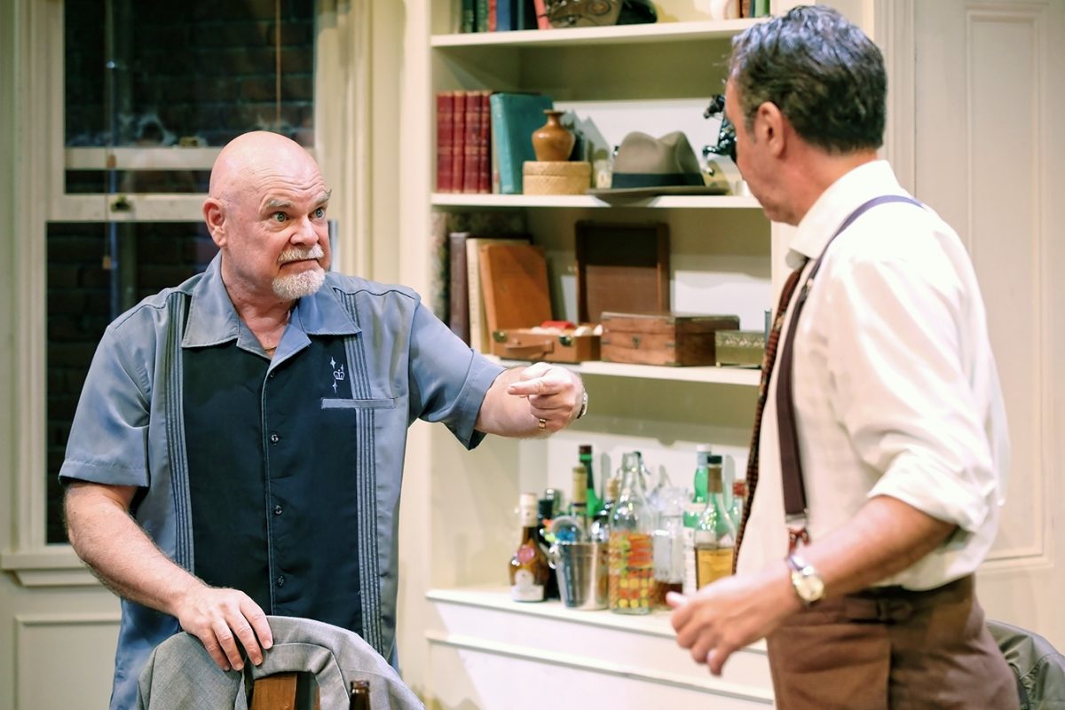 Two men have an intense conversation in a room with shelves, books, and various bottles. One man, bald and bearded, gestures while seated, facing the other man in a white shirt and braces.