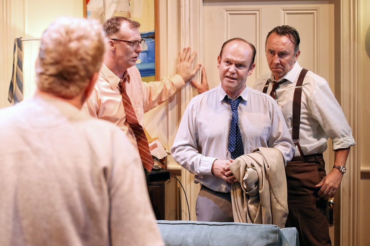 Three men in dress shirts and ties stand indoors, looking seriously at another person whose back is to the camera. One man holds a coat, and another leans against the doorframe. They appear to be in a tense conversation.