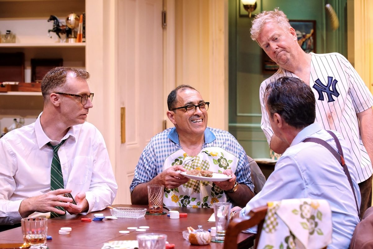 Four men sit around a table playing cards. One, wearing an apron and holding a plate of food, smiles whilst the others watch. The man standing wears a Yankees jersey. The scene is set in a cosy, brightly lit room.