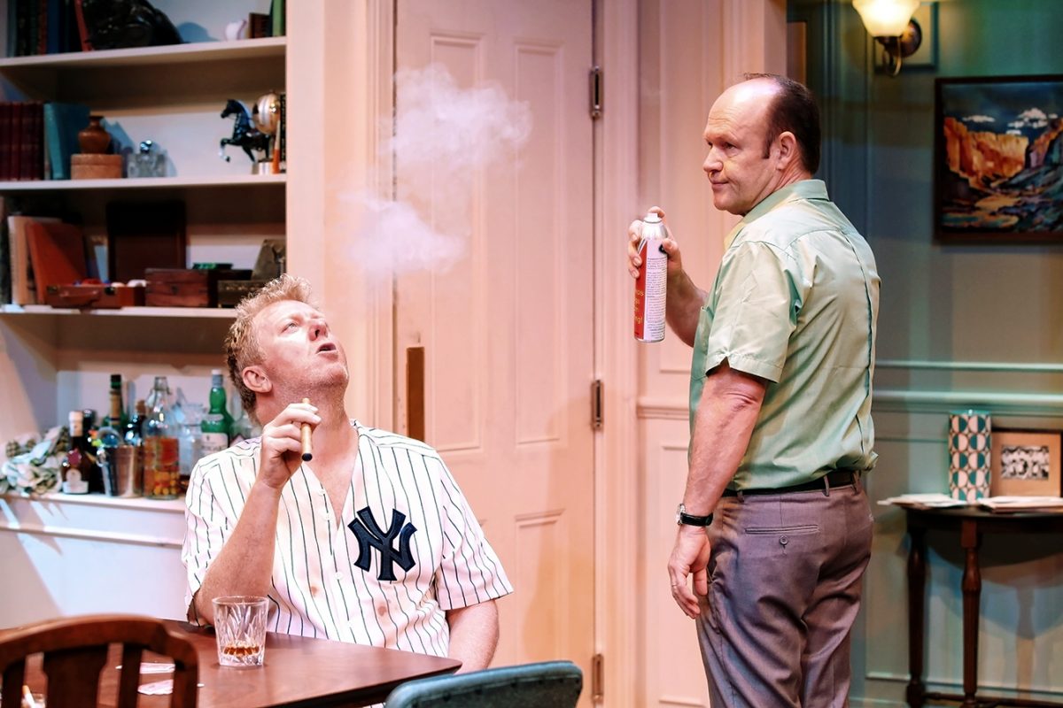 Two men are in a room; one in a Yankees jersey blows smoke upwards while seated, the other stands holding an air freshener, looking at him. The setting resembles a cosy, cluttered living room or study.