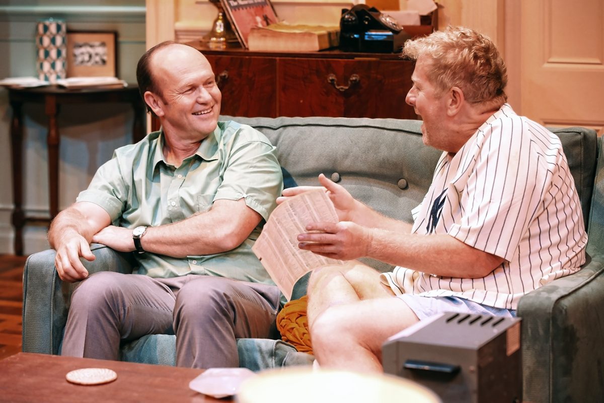 Two men sit on a sofa in a cosy living room, engaged in an animated conversation. One holds a sheet of paper and gestures enthusiastically, while the other smiles and listens attentively.
