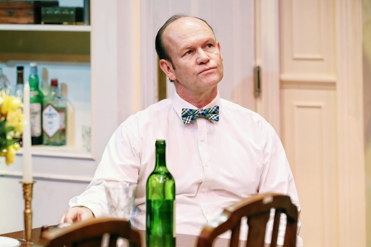 A middle-aged man in a pink shirt and checked bow tie sits at a dining table with a green wine bottle, glass, and candle, looking thoughtful. Bottles are visible on a shelf in the background.
