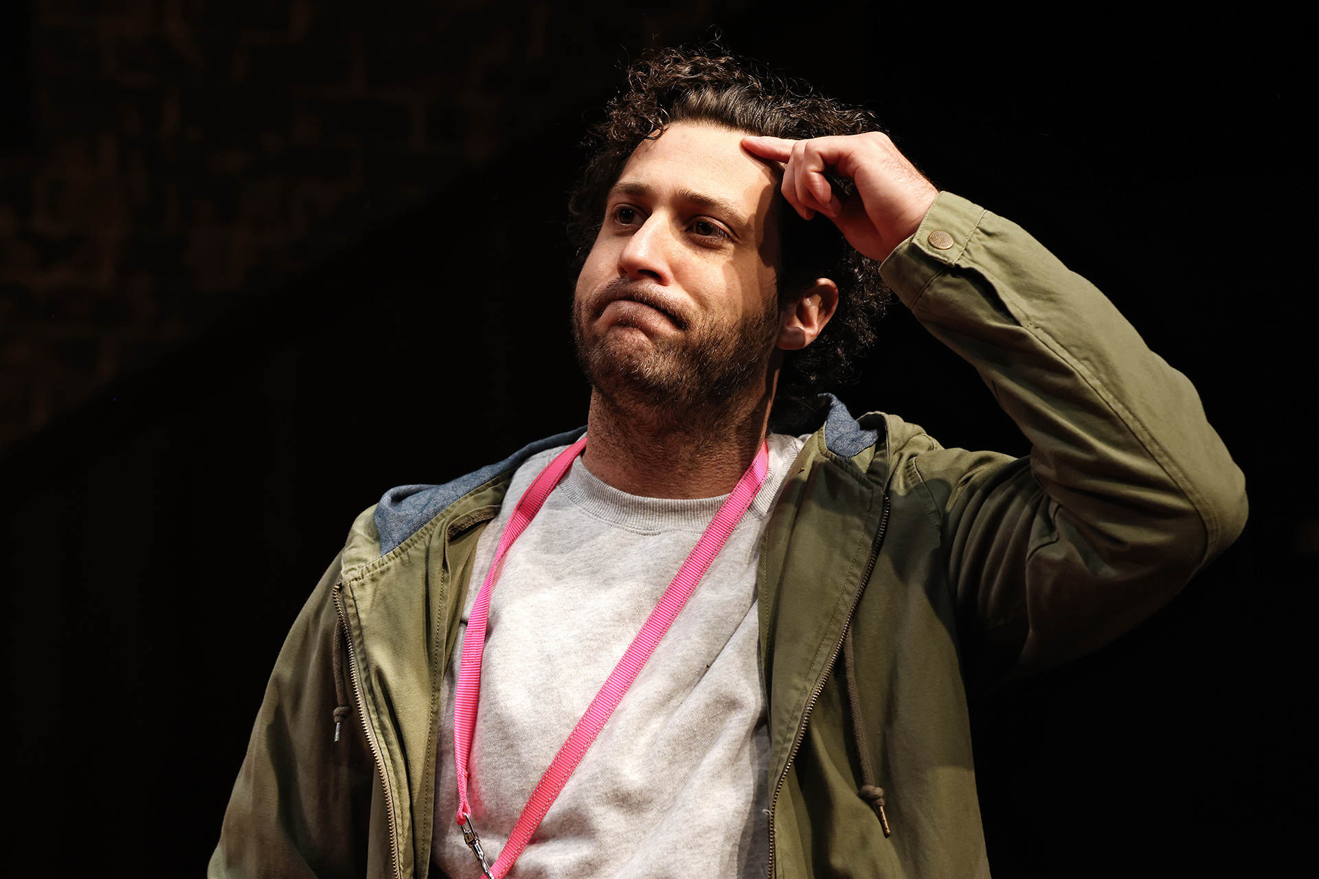 A man with curly hair and a beard stands wearing a green jacket and grey shirt. He touches his forehead with one finger, looking thoughtful or puzzled, and a pink lanyard hangs around his neck.