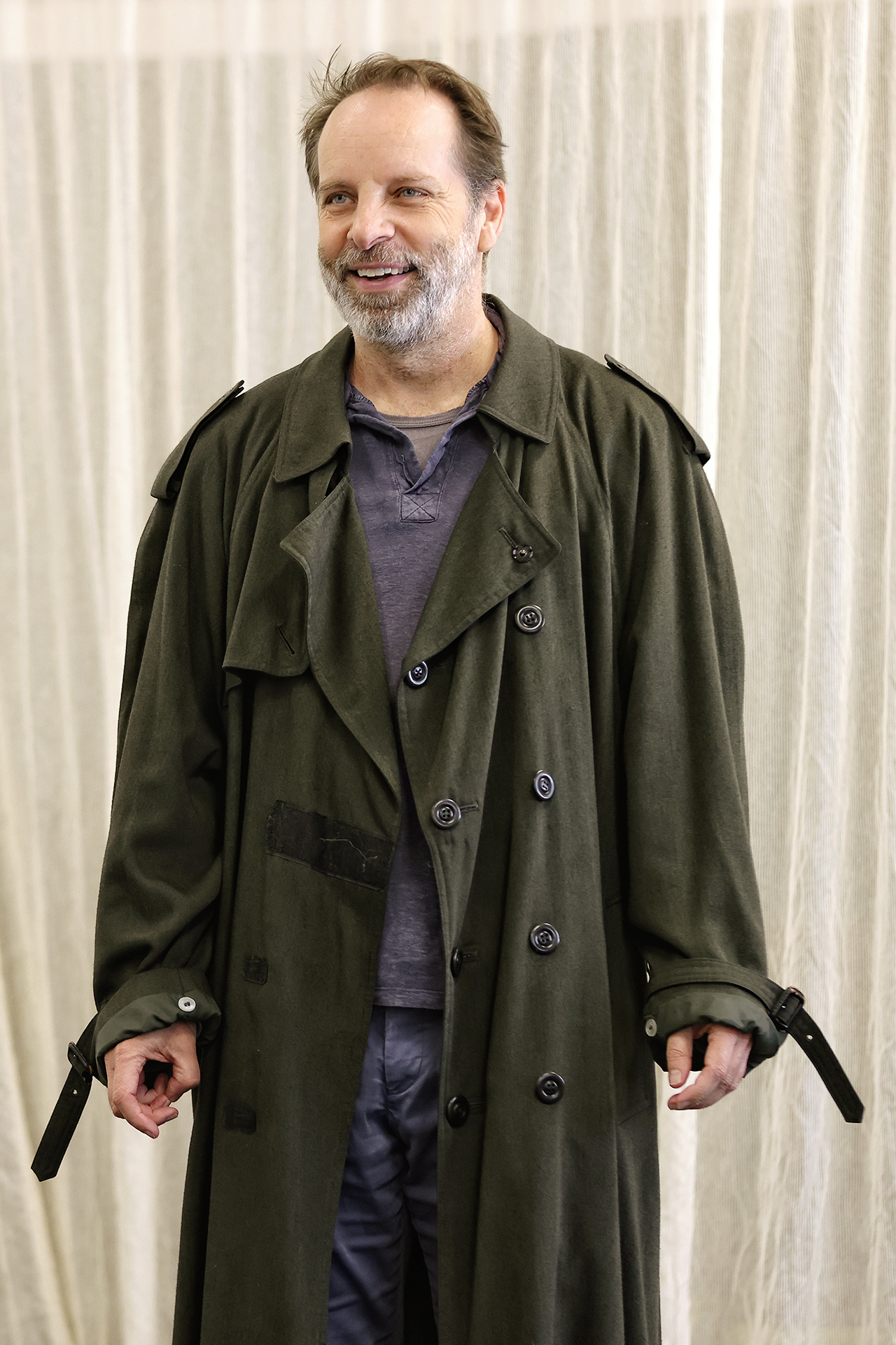 A man with a beard and moustache smiles whilst wearing a long, oversized dark trench coat over a casual shirt, standing in front of a light-coloured, textured curtain.