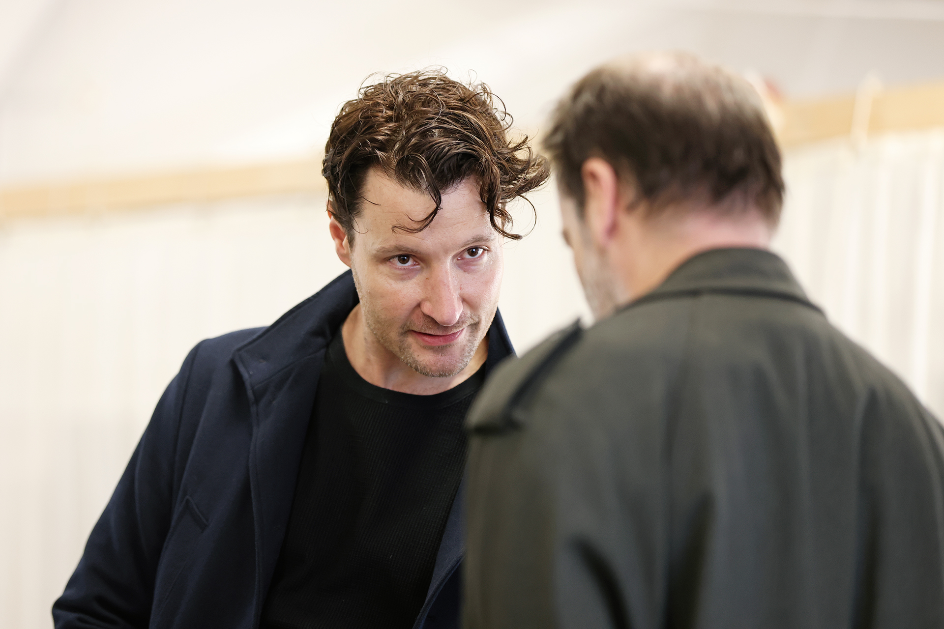 A man with curly hair and a dark coat looks intently at another man with greying hair, who is seen from behind, in a room with white curtains in the background.