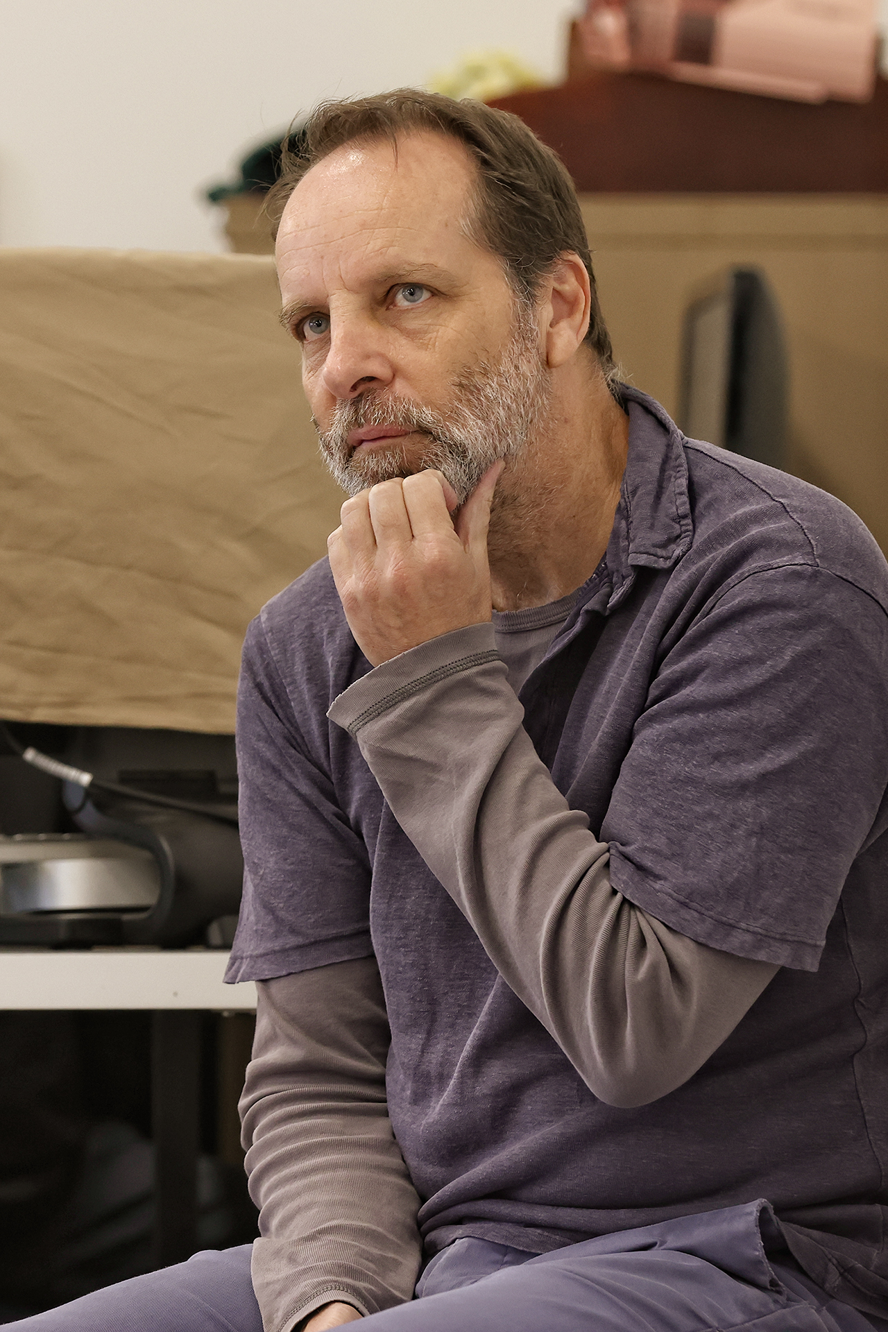 A man with a beard, wearing a purple shirt layered over a grey long-sleeve, sits indoors with his hand thoughtfully touching his chin, looking slightly upward and lost in thought.