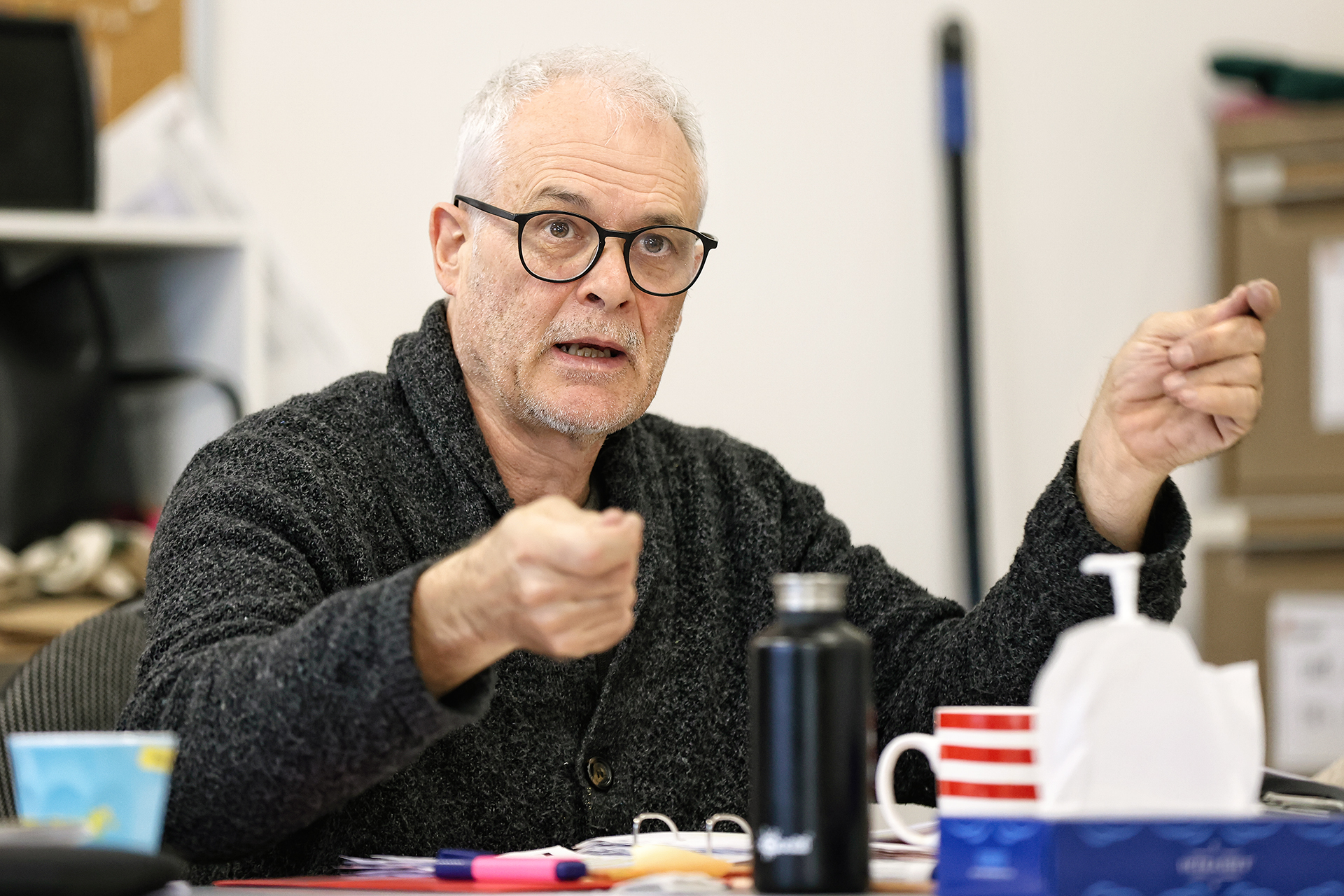 An older man with grey hair and glasses, wearing a dark jumper, gestures with both hands while speaking at a table. There are cups, a water bottle, papers, and a bottle of hand sanitiser in front of him.