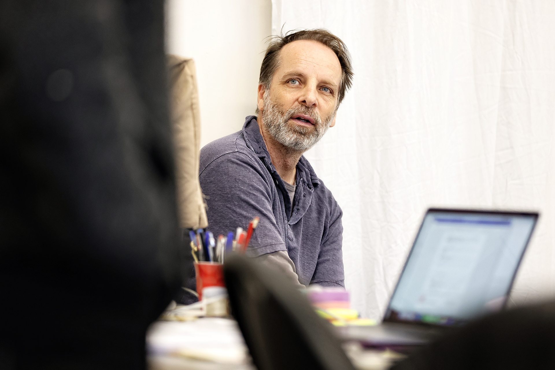A middle-aged man with a beard and moustache sits at a desk with pens and a laptop, looking to the side. The background features a white curtain, and his expression appears attentive.