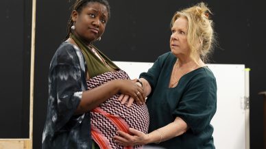 Two women stand together; one appears to be pregnant, holding her belly and looking upwards, while the other gently touches her stomach and looks at her with concern. Both wear loose, casual clothing inside a room.