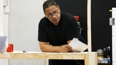 A man with glasses leans on a wooden table, holding papers and looking forward. He wears a black shirt, and a red mug and other items are on the table in a modern indoor setting.