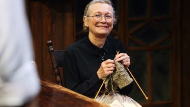 An older woman with glasses and a black blouse sits at a wooden table, smiling while knitting with brown wool. She holds knitting needles and appears content in a warmly lit, rustic room.