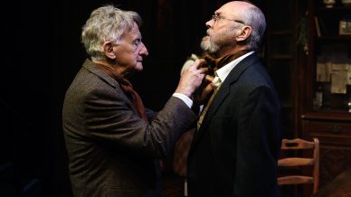 Two elderly men in suits stand face to face indoors. One man adjusts the other's tie while both look serious. They are in a dimly lit room with wooden furniture and shelves in the background.