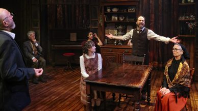 Six people in period clothing are gathered in a wooden room around a table. One man stands with arms open, appearing to speak passionately, while others sit or stand, listening intently. The atmosphere is dramatic and theatrical.