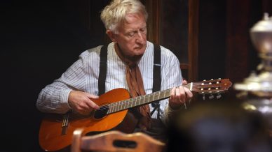 An elderly man with grey hair, wearing braces and a brown neckerchief, sits and plays an acoustic guitar indoors, focusing intently on his music.