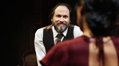 A bearded man in a waistcoat and tie sits smiling, facing a woman with dark hair in a burgundy dress, whose back is to the camera. The background is dimly lit, suggesting a theatre or stage setting.