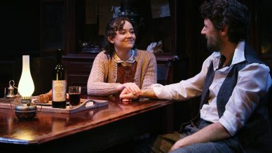 A woman and a man sit at a wooden table holding hands and smiling at each other. The table has a wine bottle, glass, lamp, and bread. The setting appears cosy and warmly lit, suggesting an intimate, domestic scene.
