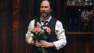 A bearded man in a waistcoat and rolled-up sleeves stands indoors, holding a bouquet of pink roses. Shelves with vases and bottles are in the background against a wooden wall.