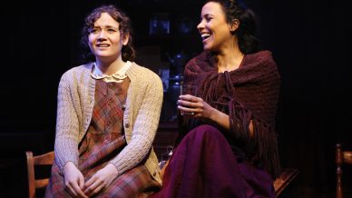 Two women sit together on stage, dressed in old-fashioned clothing. One looks concerned whilst the other smiles and holds a glass, suggesting an emotional moment in a theatrical performance.