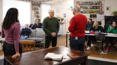 A group of people sits and watches as two older men stand facing each other in a bright classroom. A woman stands nearby with her back to the camera. Open notebooks and pens are on a wooden table in the foreground.