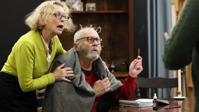 An older man with a blanket over his shoulders sits at a table, holding a pencil and looking surprised. A woman beside him, also appearing concerned, leans over to comfort him. Papers and a medicine bottle are on the table.