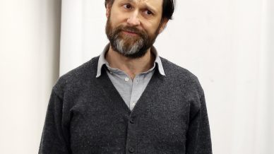 A man with a beard and moustache stands against a plain white background, wearing a dark grey cardigan over a light shirt and black trousers, looking upwards with a slightly puzzled expression.