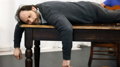 A man with a beard lies face down on a wooden table, looking tired or dejected, with his arms hanging limply off the sides. A person in the background plays a guitar.