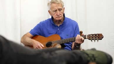 An older man in a blue polo shirt sits and plays an acoustic guitar, looking thoughtful. The foreground is blurred, showing a person lying down with green trainers. The background features a white curtain.