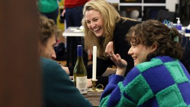 Three people sit at a table talking and laughing. A woman with blonde hair smiles widely, whilst another woman in a green and blue jumper gestures. A wine bottle and unlit candle are on the table. The background shows a busy room.