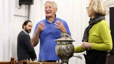 An older man in a blue polo shirt gestures and smiles whilst speaking to a woman in glasses and a green jumper beside a large metal samovar. Another man in grey stands in the background, observing.