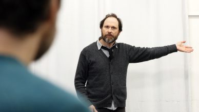 A man with a beard and messy hair stands in front of a white curtain, wearing a grey cardigan and tie, gesturing with his right arm extended. Another person is visible out of focus in the foreground.