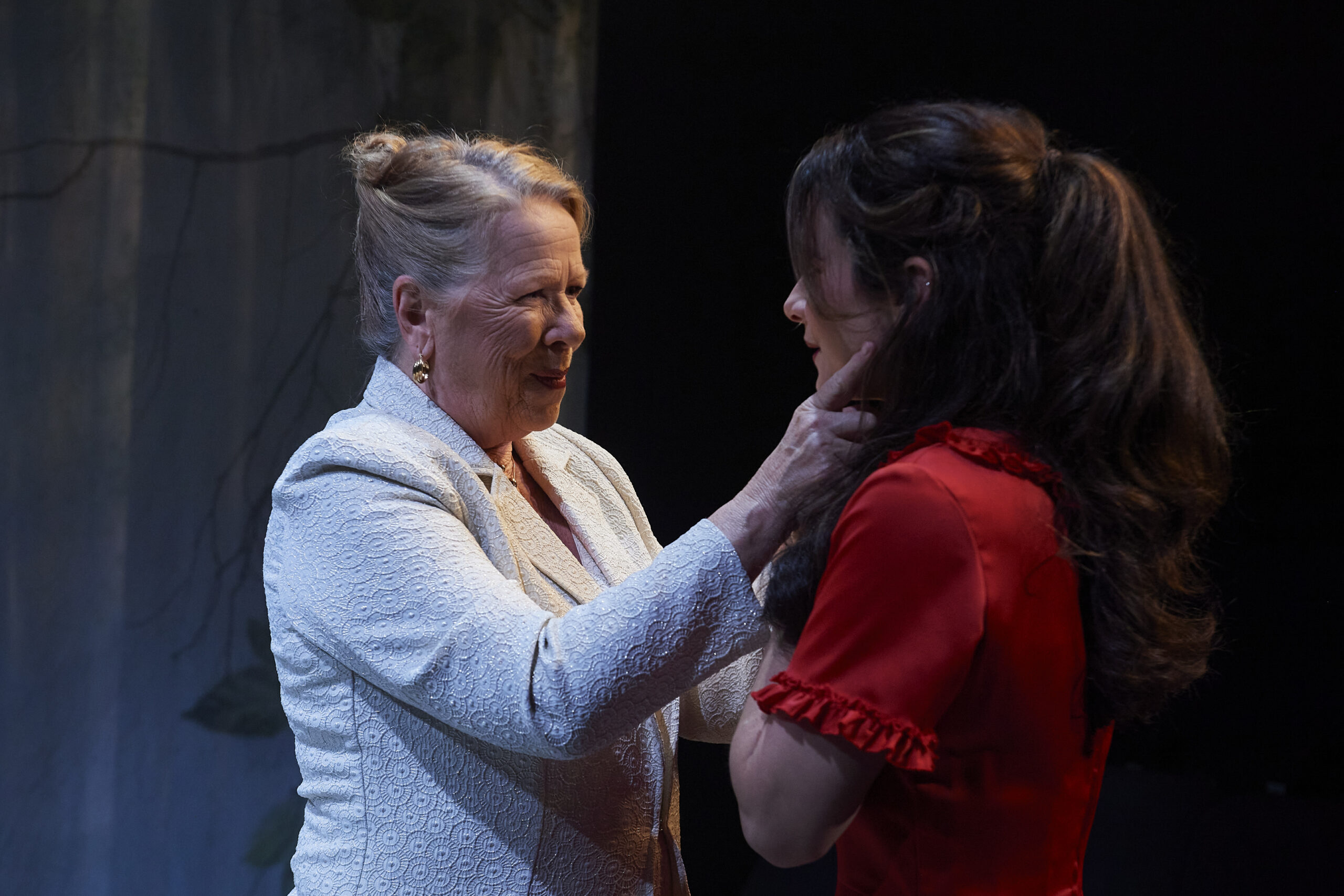 An older woman in a white blazer gently holds the face of a younger woman in a red dress, both sharing an emotional moment on a dimly lit stage.