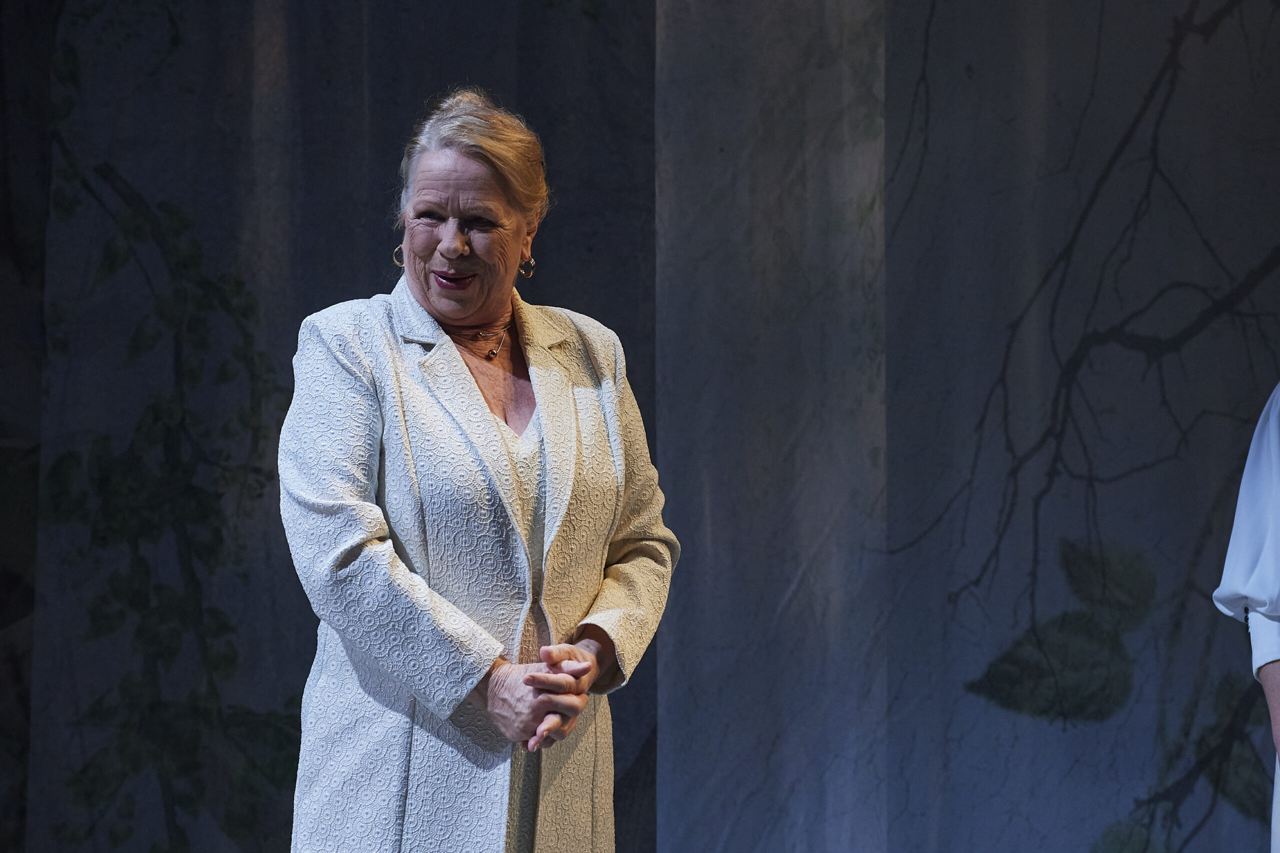 An older woman in a light patterned coat stands on stage, smiling with her hands clasped. The background is softly lit with tree branch shadows.