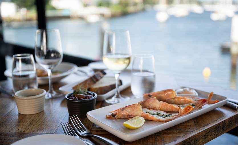 A plate of grilled prawns with a lemon wedge is set on a wooden table with cutlery, wine glasses, water glasses, and small dishes, overlooking a waterfront view.