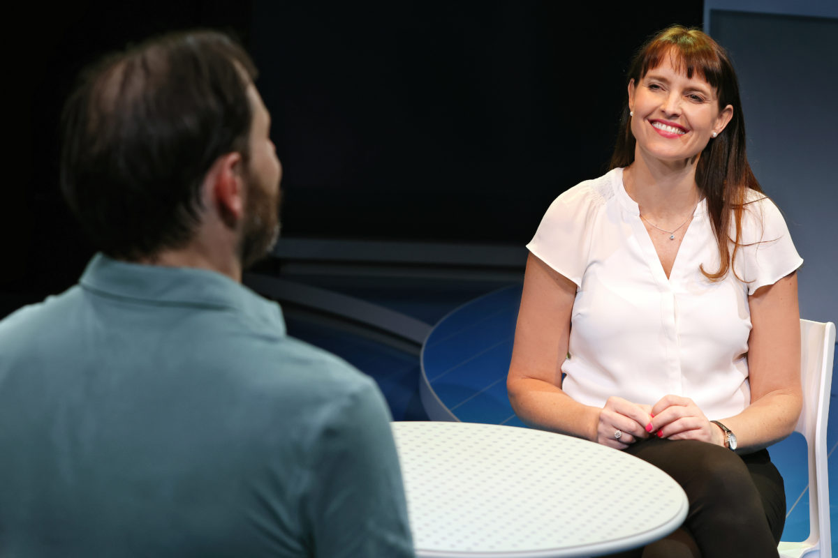 A woman in a white blouse smiles whilst sitting at a round table opposite a man in a teal shirt. They appear to be having a friendly conversation in a well-lit indoor setting.